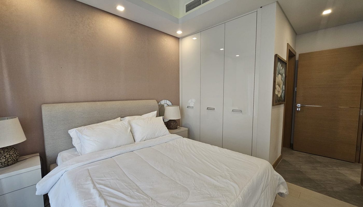 Modern bedroom with a neatly made bed, white linens, nightstands with lamps, closed white wardrobes, and a wooden door. Walls are painted light tones, and the room is well-lit.