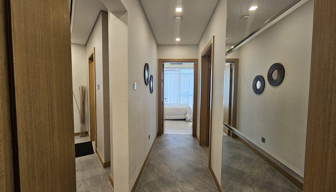 Hallway with wooden doors, a mirrored wall on the right, gray tiled floor, decorative wall hangings, and a view leading to a well-lit bedroom.