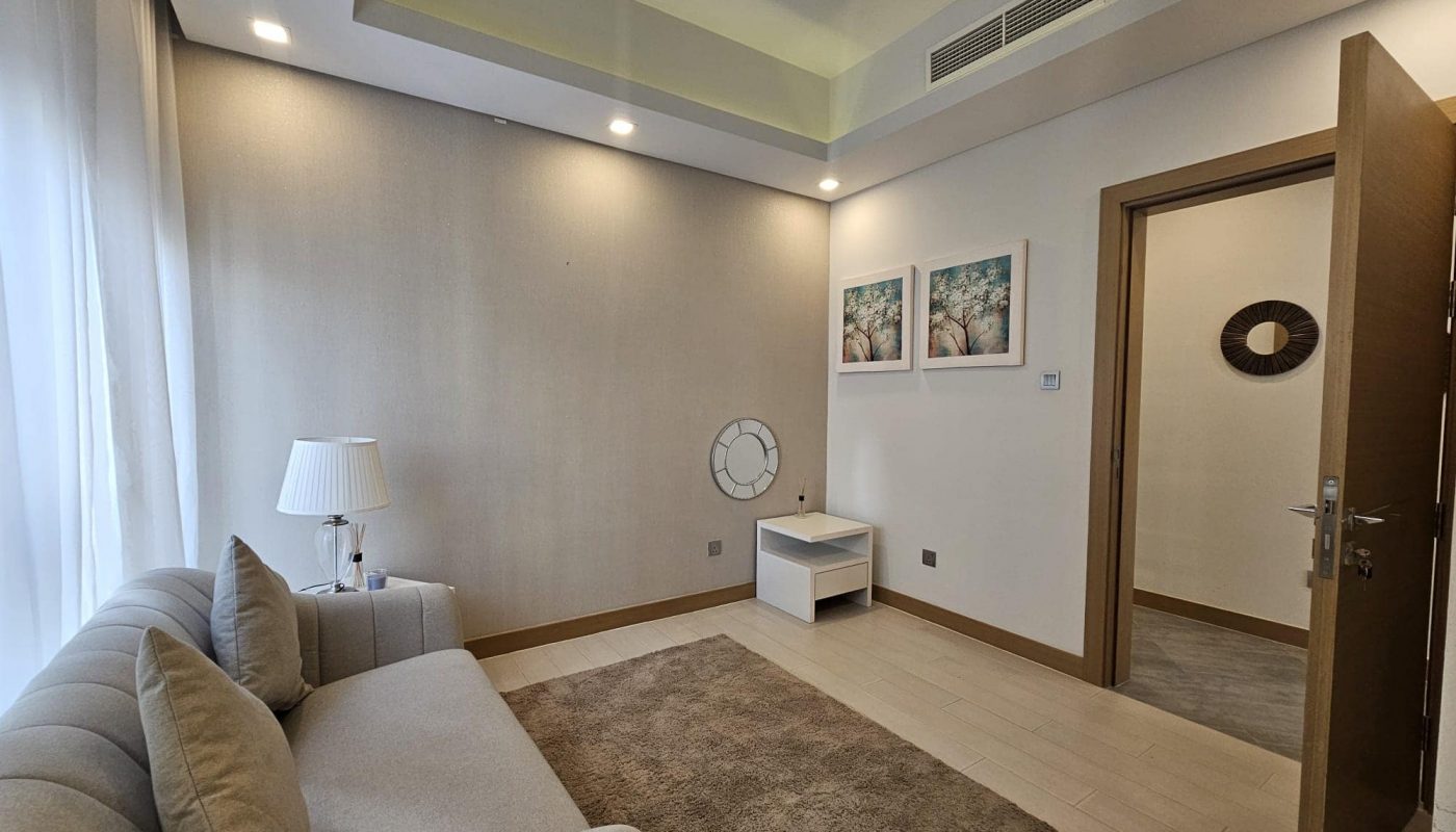 A small room with a gray sofa, beige rug, and white side table. Two artworks are on the wall, and a lamp is beside the sofa. A door is partially open, and there's a mirror on the hallway wall.