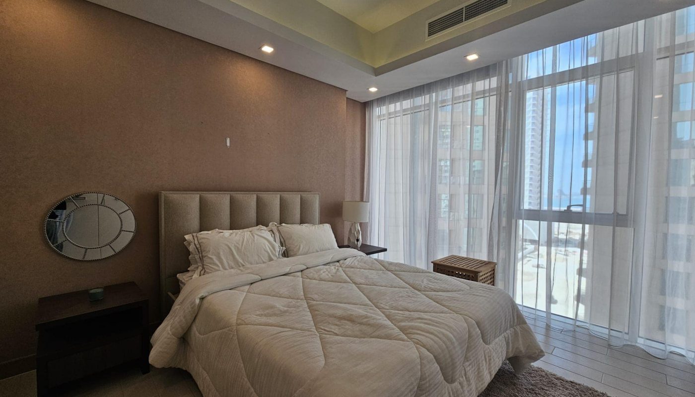 A neatly made bed with white linens in a modern bedroom. Large windows with sheer curtains allow natural light to fill the room. A small round mirror and bedside tables are visible.