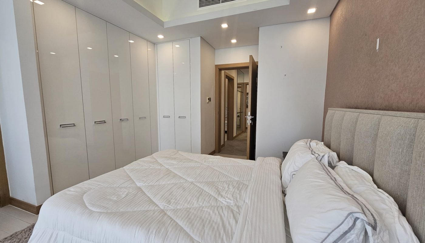 A modern bedroom with a beige upholstered bed, white bedding, built-in wardrobes, recessed lighting, and a carpeted floor. A door leads to an adjacent room.