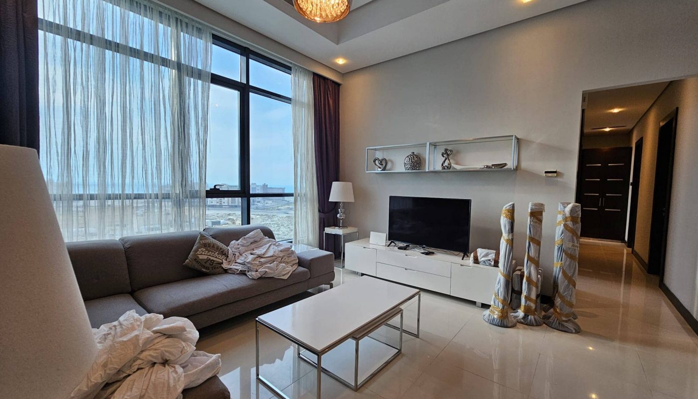 Modern living room interior in a fully-furnished 2 bedroom apartment with ocean view through large window, featuring a beige sofa, white tv cabinet, and glass coffee table. For Rent.