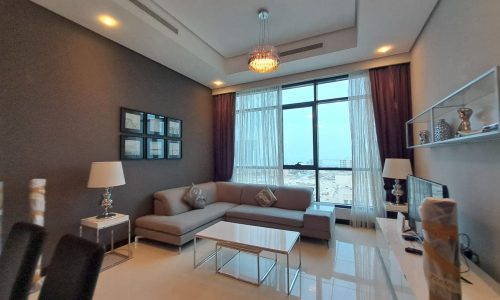 Modern living room in a fully-furnished 2 bedroom apartment with gray sofas, glass coffee table, maroon curtains, and a view of the city through large windows. Decor includes lamps and a wall