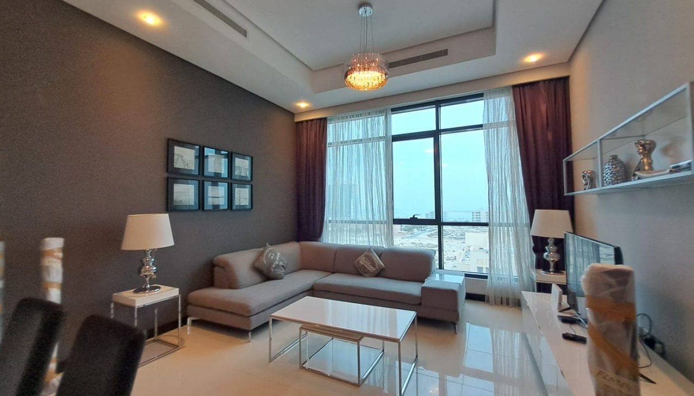 Modern living room in a fully-furnished 2 bedroom apartment with gray sofas, glass coffee table, maroon curtains, and a view of the city through large windows. Decor includes lamps and a wall