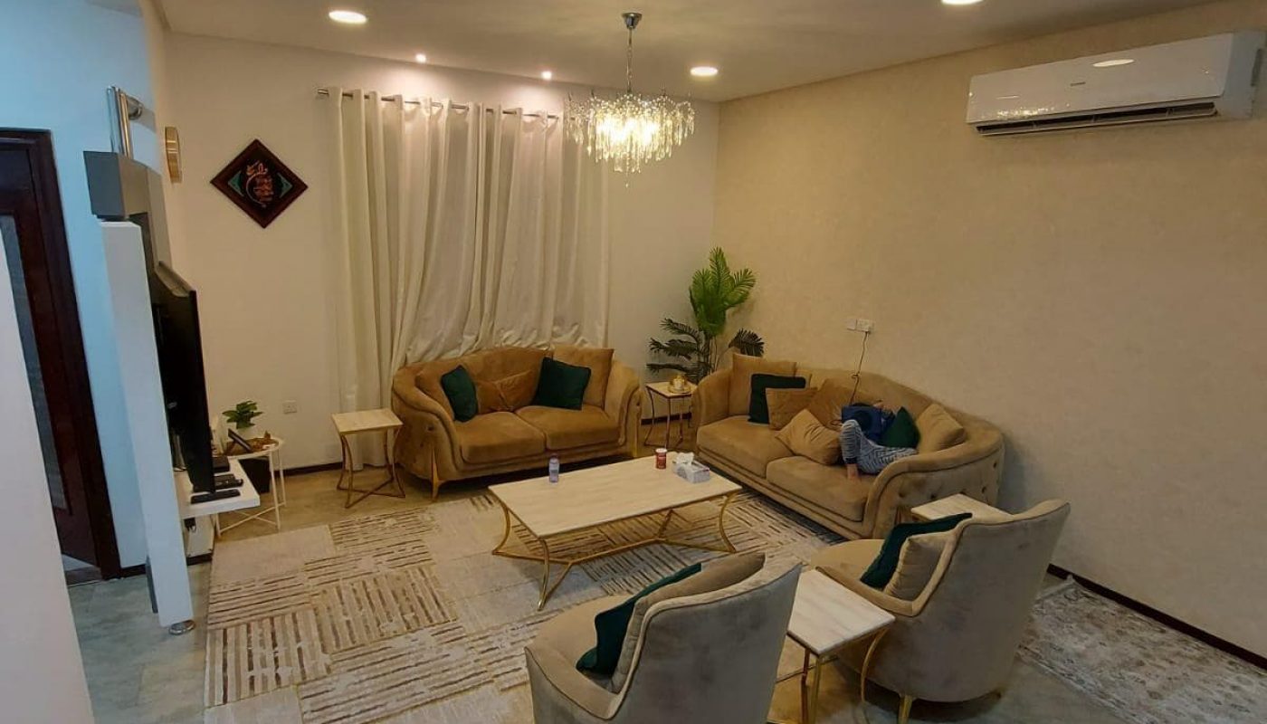 A modern living room in a Family 3BR Villa with cream walls featuring two beige sofas, two gray armchairs, a chandelier, and an air conditioning unit.
