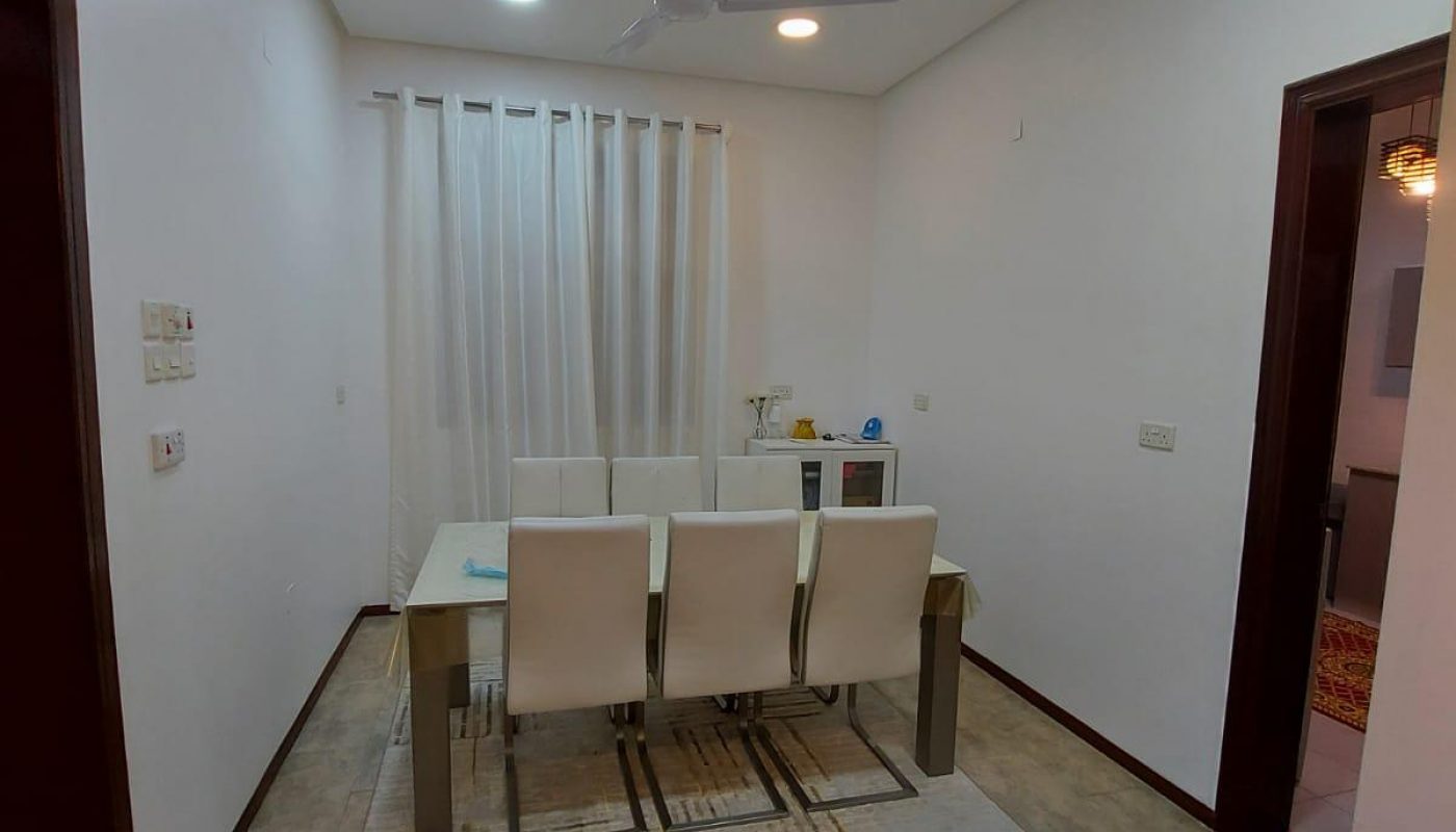 A modern dining room in a 3BR villa with a white table and chairs on a textured rug, featuring bright ceiling lights and sheer curtains.
