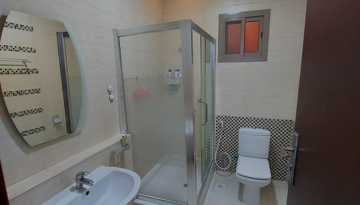 A modern bathroom in a family 3BR villa with a shower stall, toilet, sink, and a round mirror on a tiled wall, illuminated by an overhead light.