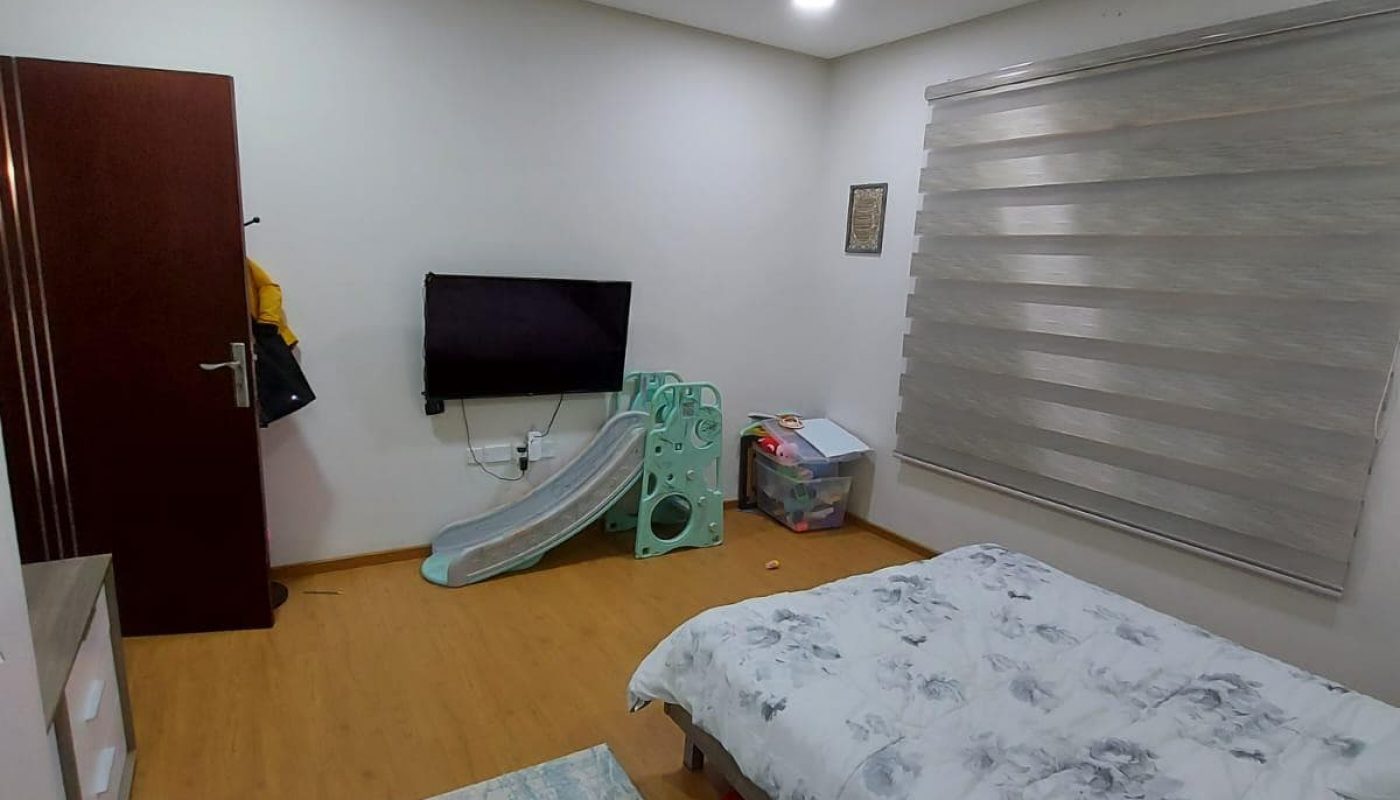 A tidy bedroom in a Family 3BR Villa with a bed, wall-mounted TV, ceiling fan, and a sliding window blind. An ironing board and various items are in one corner.