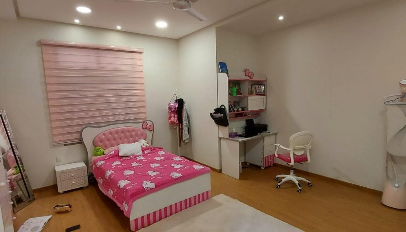 A tidy bedroom in a family 3BR villa with a single bed covered in a pink floral duvet, a study area with a desk and chair, a large white rug on the floor, and