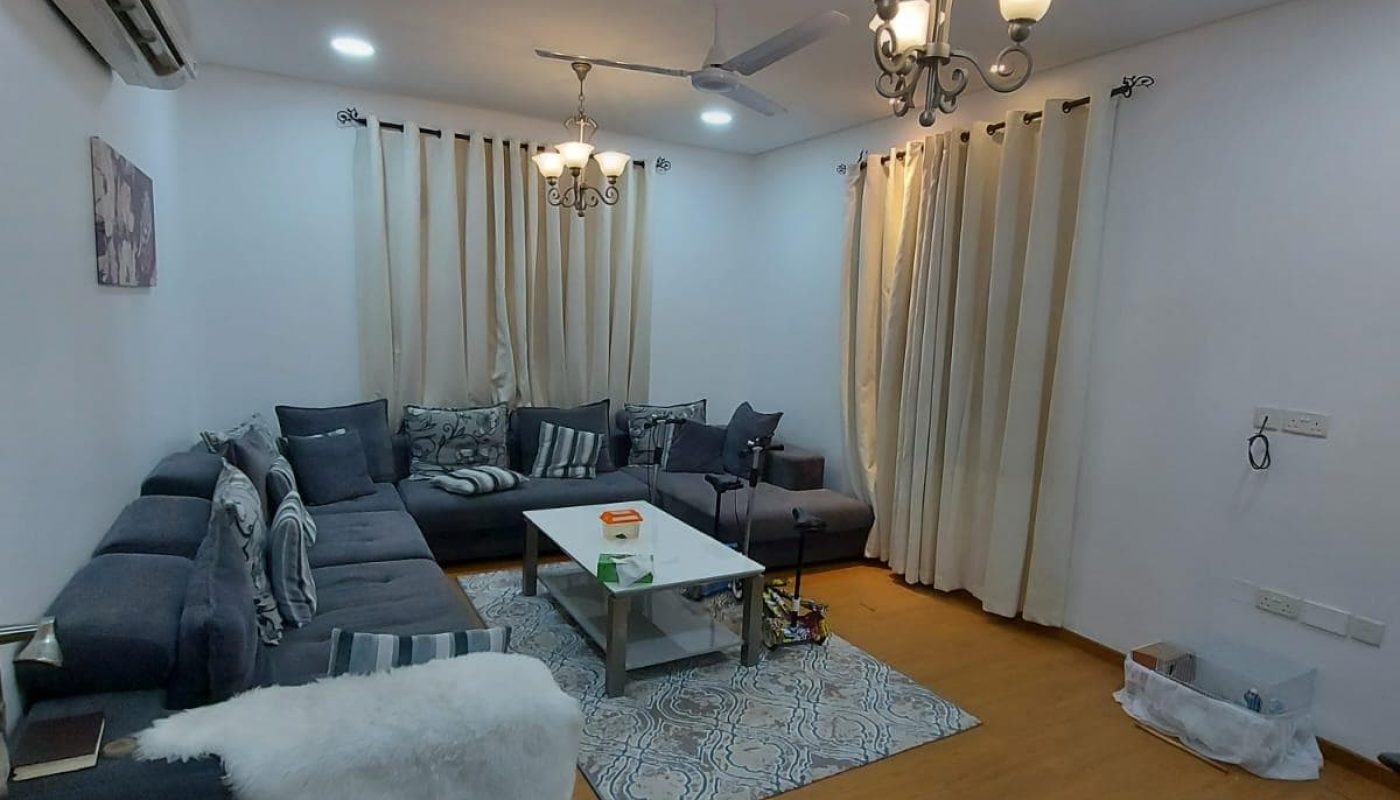 Modern living room in a family 3BR villa with a large blue sectional sofa, beige curtains, white walls, and a central coffee table. A bicycle is parked on the wooden floor near the entrance