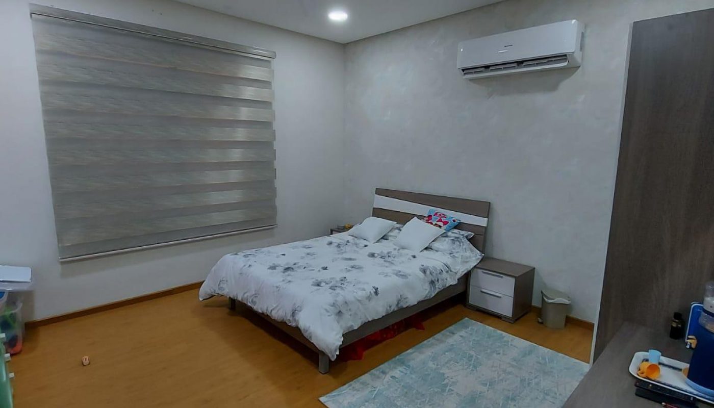 Modern 3BR family villa for sale in Jid Ali Area, featuring a bedroom with a double bed, white and grey bedding, an air conditioner, ceiling fan, wooden flooring, and a small