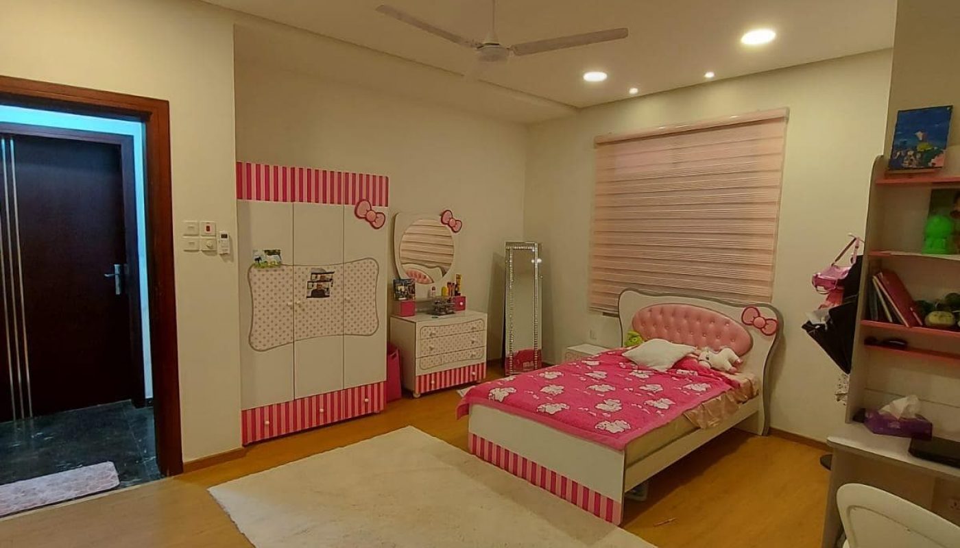 A tidy child's bedroom, within a family 3BR villa for sale in the Jid Ali area, featuring a pink-striped wall, a single bed, and various toys. A plush white rug