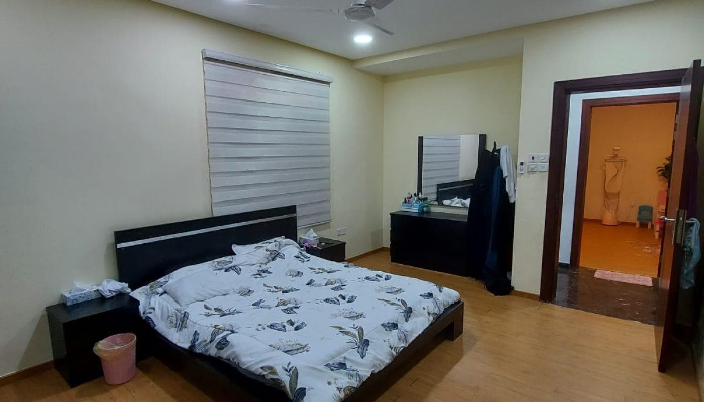 A well-lit bedroom in a family 3BR villa for sale, featuring a double bed, patterned bedding, a ceiling fan, wooden floor, and an open door leading to another room.