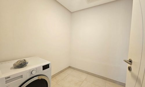 A small, empty laundry room with a white washing machine, beige tiled floor, and plain white walls. an open door shows on the right.