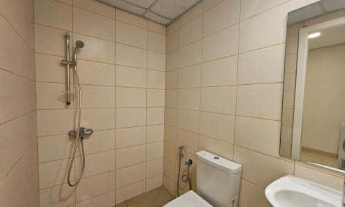 A small bathroom with beige tiles, featuring a shower, toilet, and sink, with visible mold on the ceiling corner.