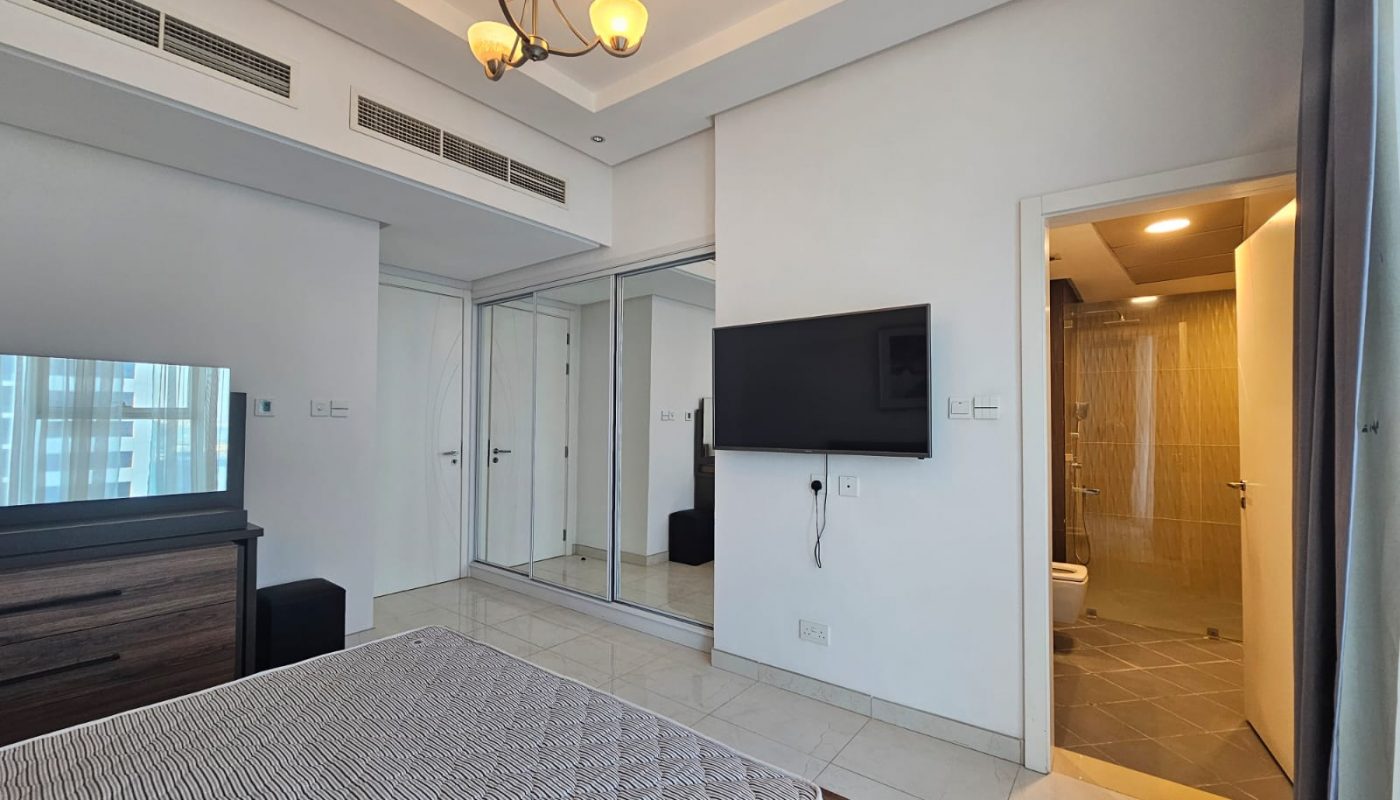 Modern flat in Juffair with a large bed, mounted TV, mirrored closets, and an open door leading to a bathroom. Brightly lit with natural light and elegant lighting fixtures.