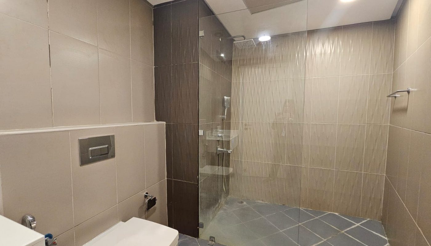 Modern flat bathroom interior with beige tiles, a glass shower enclosure, and a toilet on the left side.