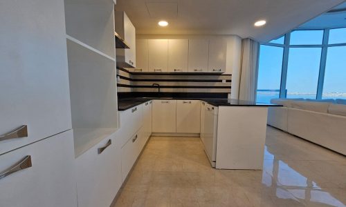 Modern kitchen with white cabinets, black countertop, and an open layout leading into a living room with large windows displaying an ocean view.