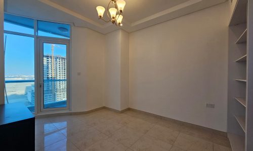 Empty room with beige floor tiles, white walls, a hanging chandelier, built-in shelves, and large windows showing a city skyline.