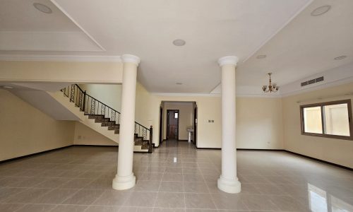 Empty living room in a villa for rent with two white columns, tiled floor, a staircase on the left, and multiple windows with natural light.