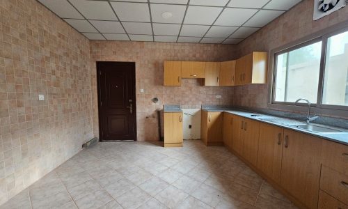 An empty kitchen in a villa for rent with beige tile flooring, wooden cabinets, and built-in sink and dishwasher, all under natural light from a window.