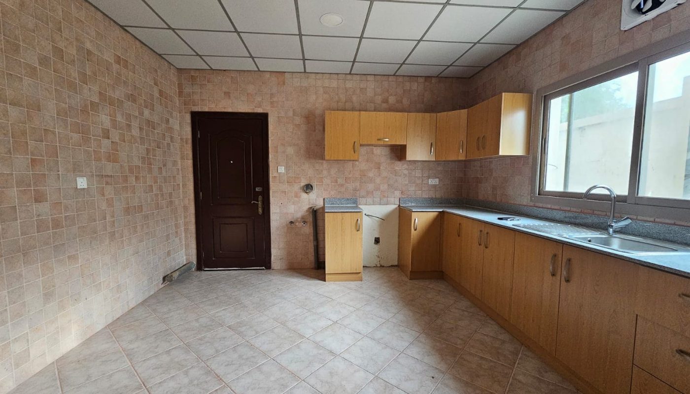 An empty kitchen in a villa for rent with beige tile flooring, wooden cabinets, and built-in sink and dishwasher, all under natural light from a window.