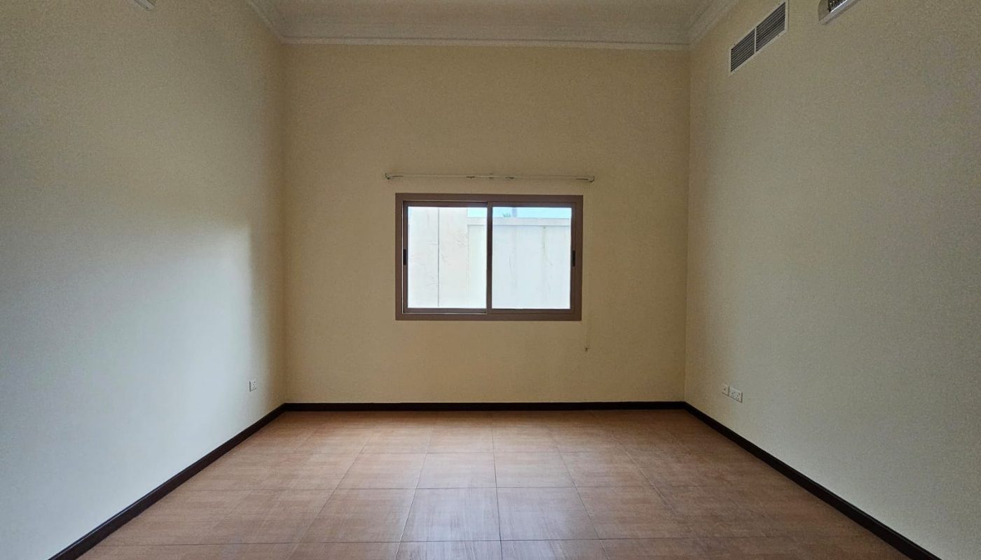 Empty room in Villa Juffair with cream walls, a single window, and wooden flooring, illuminated by natural light.