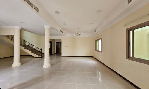 Spacious empty room in a villa for rent with polished tile flooring, two white columns, large windows, and a staircase on the right.