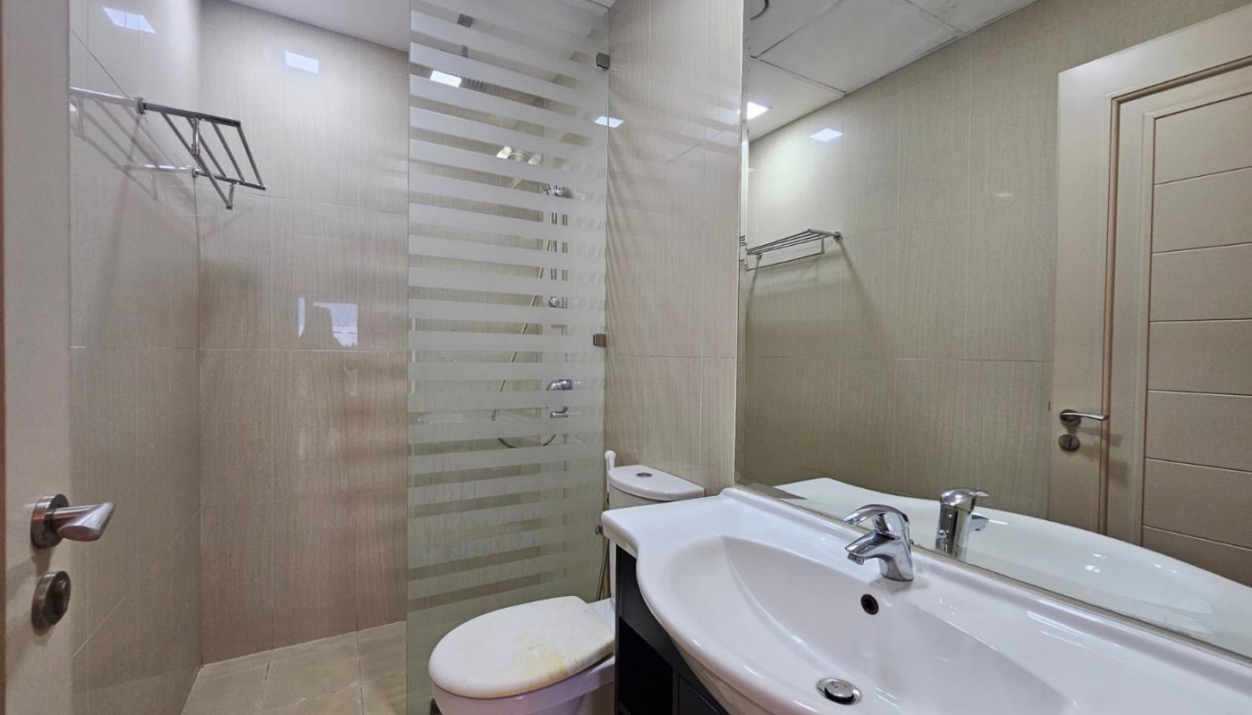 Modern apartment bathroom interior featuring a glass shower, beige tiles, white porcelain toilet and sink, and a mirror above the sink.