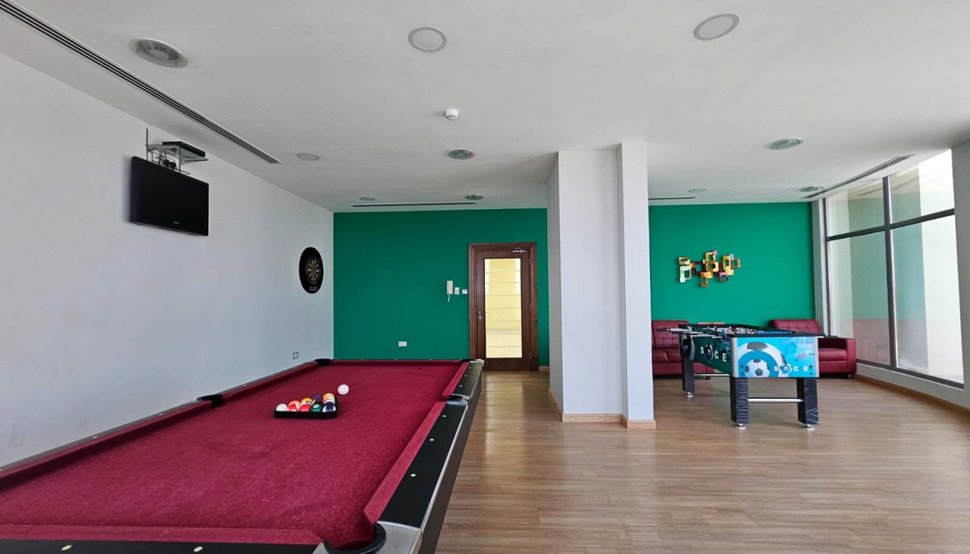 A modern apartment with a recreation room featuring a red pool table, dartboard, foosball table, wooden flooring, and a green accent wall.