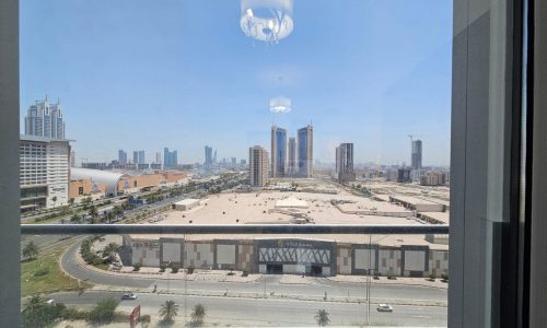 View from a window looking out on a sunny cityscape in Juffair with skyscrapers under construction, roads, and a parking lot, with a hanging lamp visible indoors.