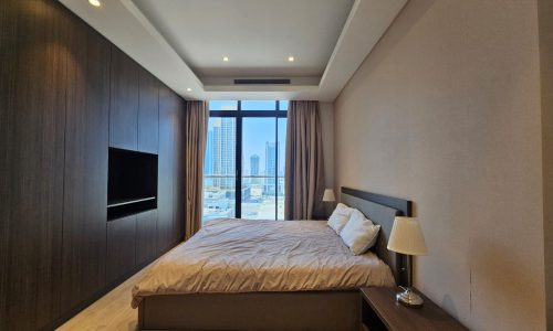 Modern bedroom with a large bed, wooden furniture, mounted tv, and a window with a city view.