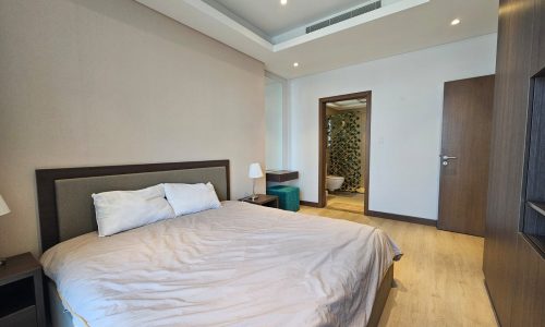 A modern bedroom with a neatly made bed, wooden floor, ceiling lights, and a view into an adjoining room with a green plant.