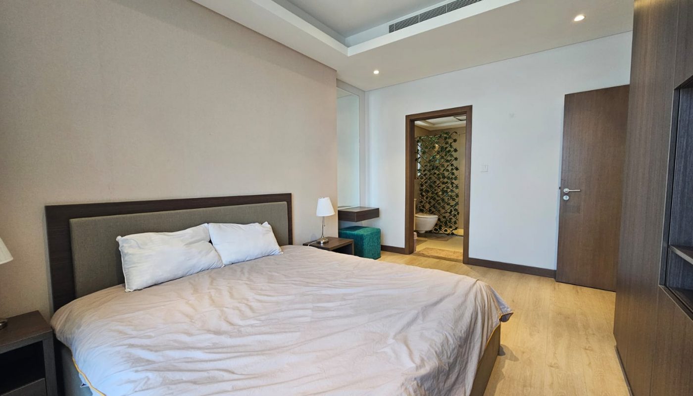 A modern bedroom with a neatly made bed, wooden floor, ceiling lights, and a view into an adjoining room with a green plant.