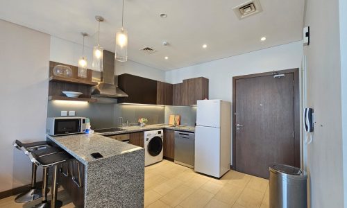 Modern kitchen interior in a Seef flat for rent featuring dark wood cabinets, stainless steel appliances, a white refrigerator, and pendant lights, with a central island.