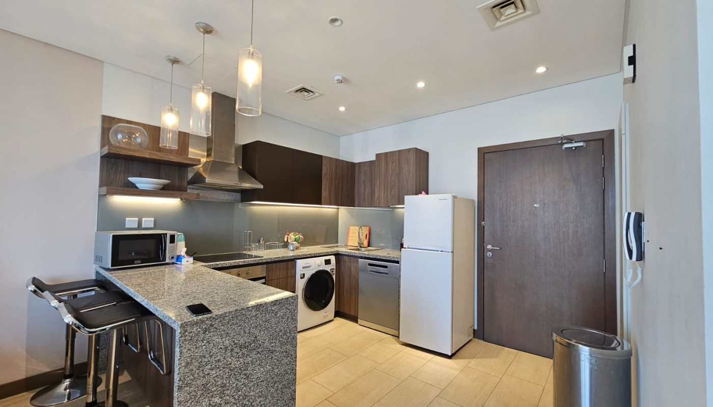 Modern kitchen interior in a Seef flat for rent featuring dark wood cabinets, stainless steel appliances, a white refrigerator, and pendant lights, with a central island.
