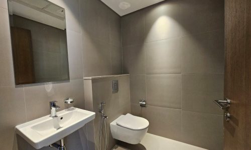 Modern bathroom in a Seef flat for rent with gray tiles featuring a wall-mounted sink, toilet, bidet spray, mirror, and flush-mounted light.