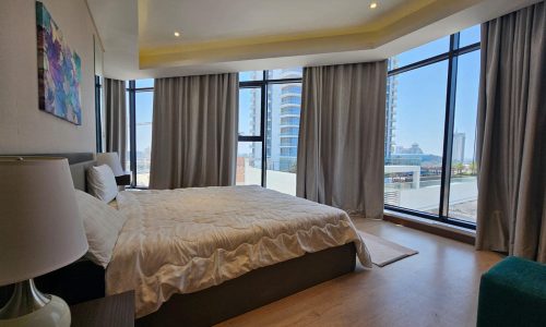 Modern flat with a large bed, floor-to-ceiling windows providing Seef city views, minimalist decor, and an abstract wall painting.