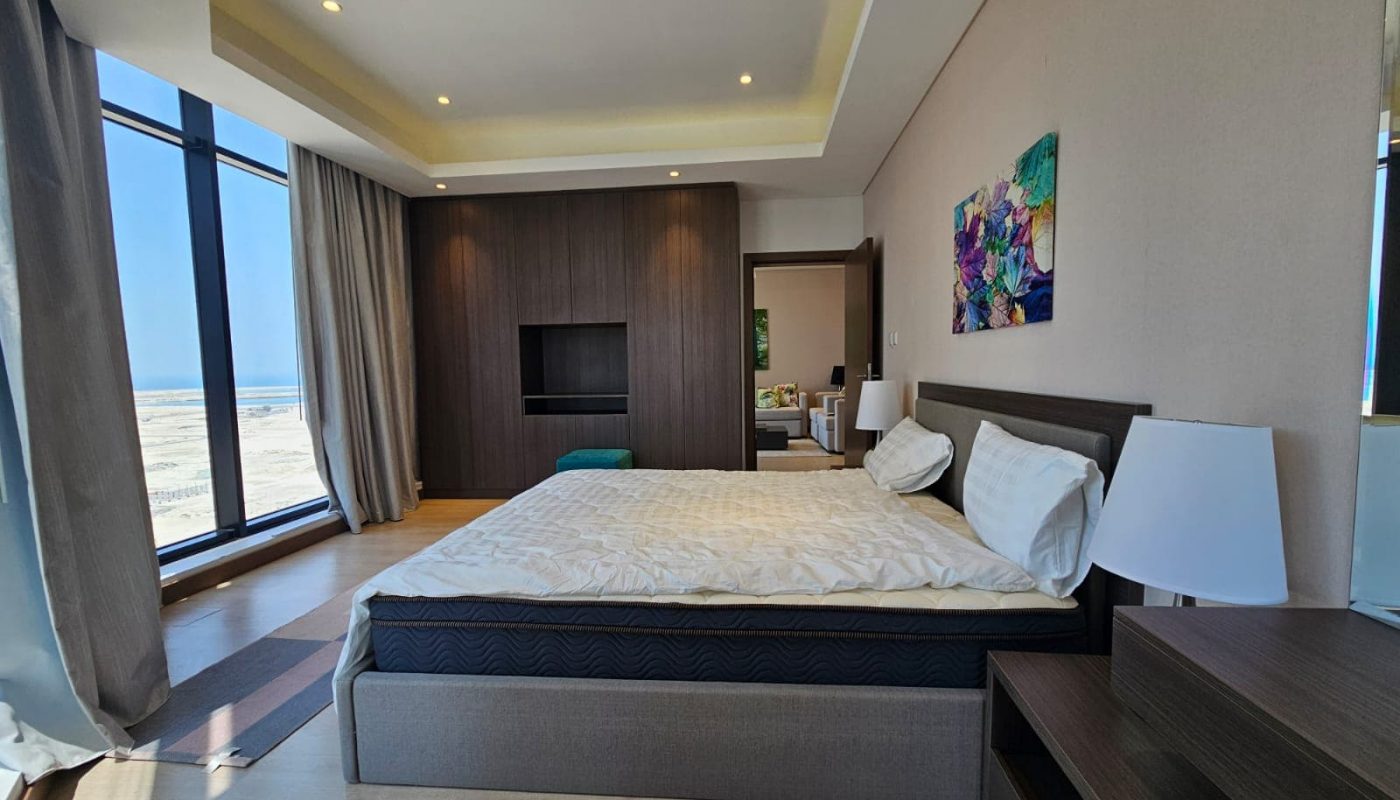 Modern flat with a large bed, wooden furniture, ceiling lights, and a seascape visible through floor-to-ceiling windows.