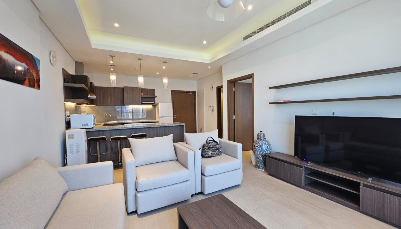 Modern flat for rent with a living room featuring white sofas, a large TV, and an integrated kitchen area, complete with sleek wooden cabinetry and ambient lighting in Seef.
