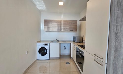 Small modern kitchen with white cabinets, washing machine, and built-in appliances, featuring tile flooring and neutral colors in a modern flat for rent.