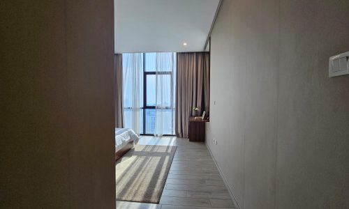 Interior of a modern flat in Juffair with a large window, partial view of a bed, and a small desk with a mirror along a clean, minimalist hallway.