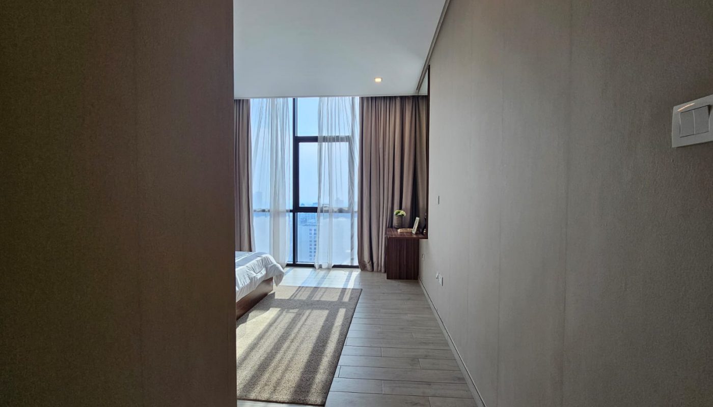 Interior of a modern flat in Juffair with a large window, partial view of a bed, and a small desk with a mirror along a clean, minimalist hallway.