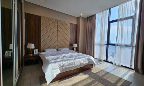 Modern flat in Juffair with a large bed centered between draped windows showcasing city views, wooden paneling, and minimalistic decor.