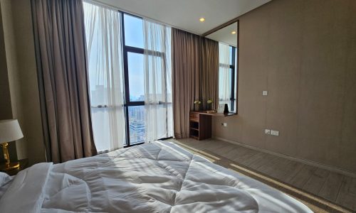 Modern flat with a large bed, sheer curtains, and a waterfront view through floor-to-ceiling windows.