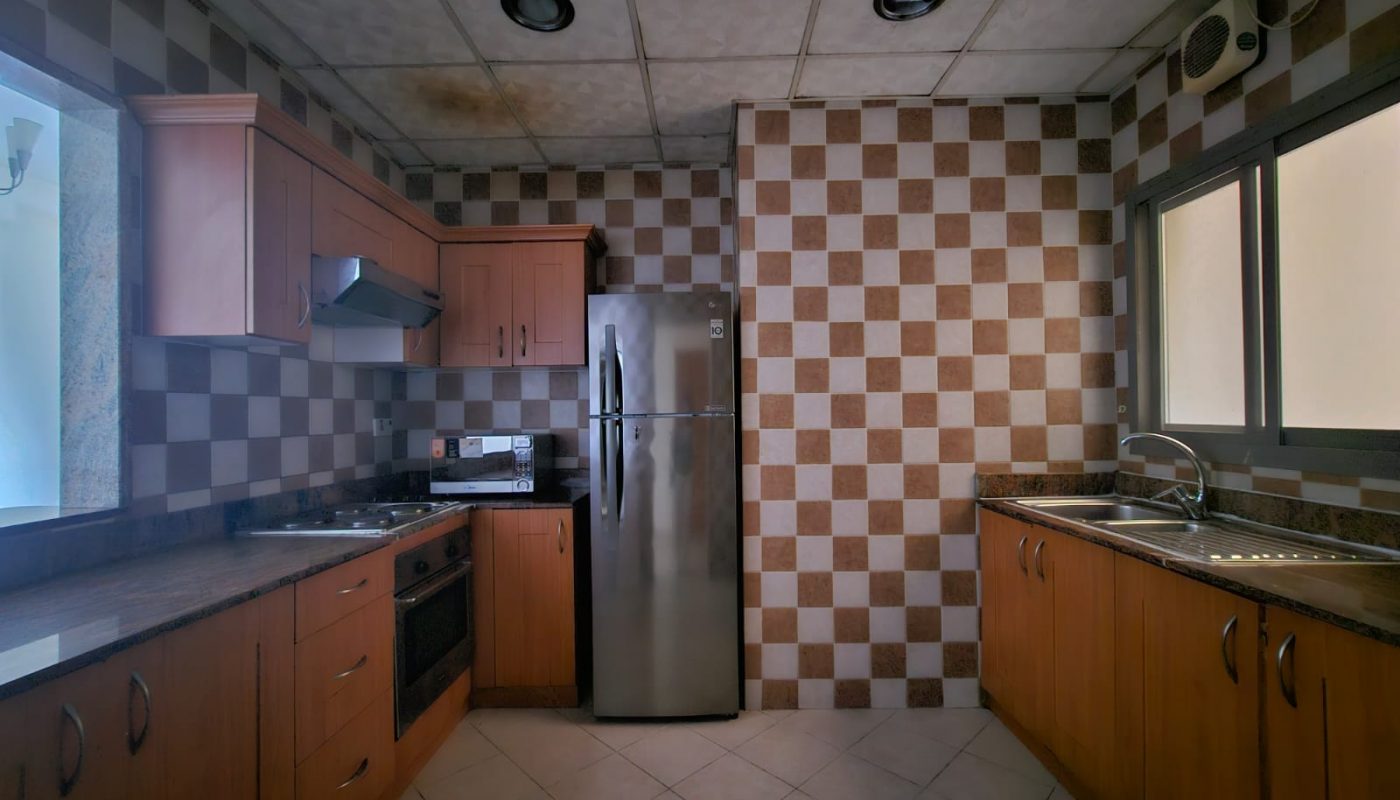 A kitchen interior featuring wooden cabinets, a stainless steel refrigerator, checkerboard wall tiles, and ample counter space in Seqya.