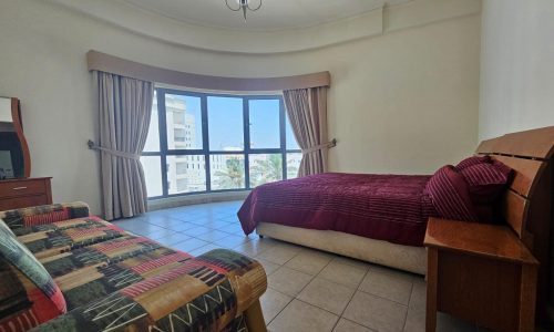 A bright, spacious bedroom in a flat for rent with two beds, striped bedding, and a large window offering a view of the ocean.