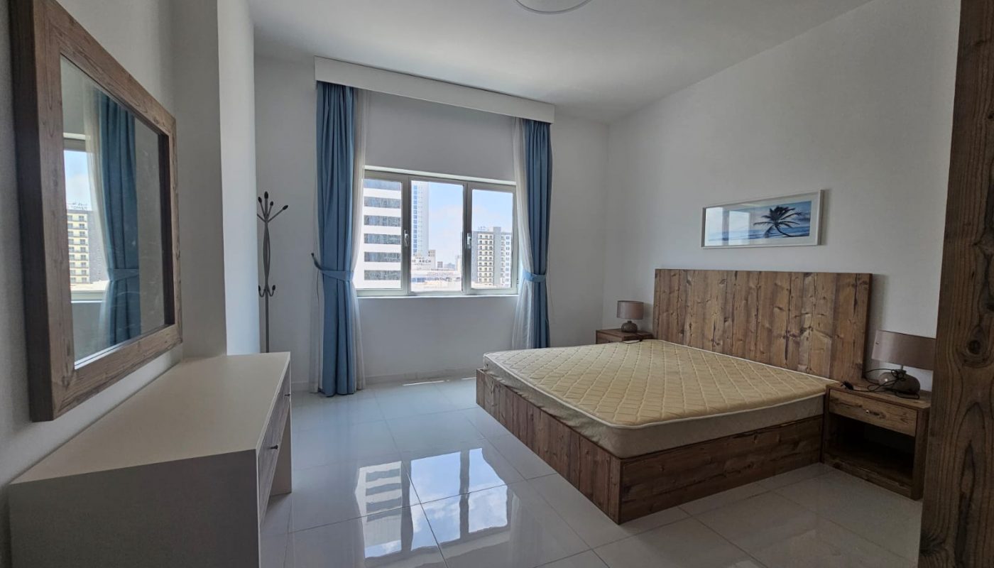 A modern two-bedroom apartment in Juffair with a wooden bed and headboard, white walls, floor tiles, and large windows with city views.