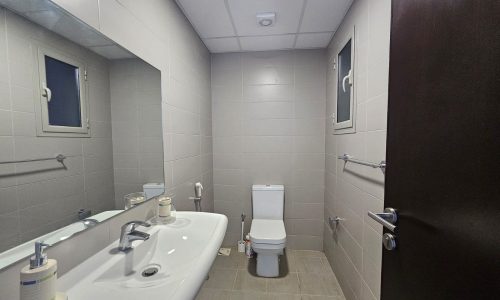 A clean, modern bathroom in a fully furnished apartment featuring two sinks with mirrors, a toilet, and gray wall tiles.