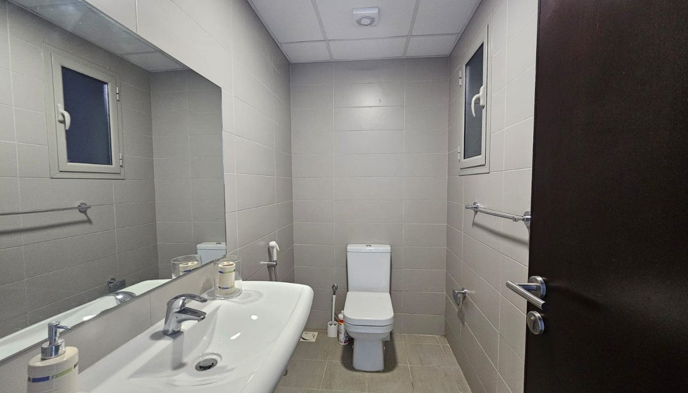 A clean, modern bathroom in a fully furnished apartment featuring two sinks with mirrors, a toilet, and gray wall tiles.